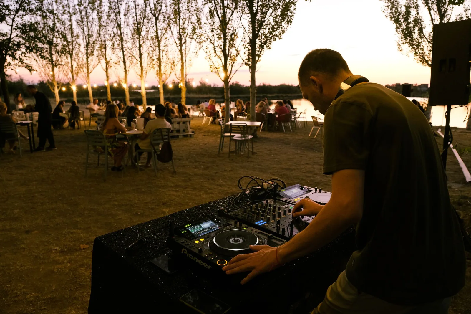 DJ tocando em mesa de som ao ar livre, à beira de um lago ao entardecer (foto de arquivo, não é do evento)