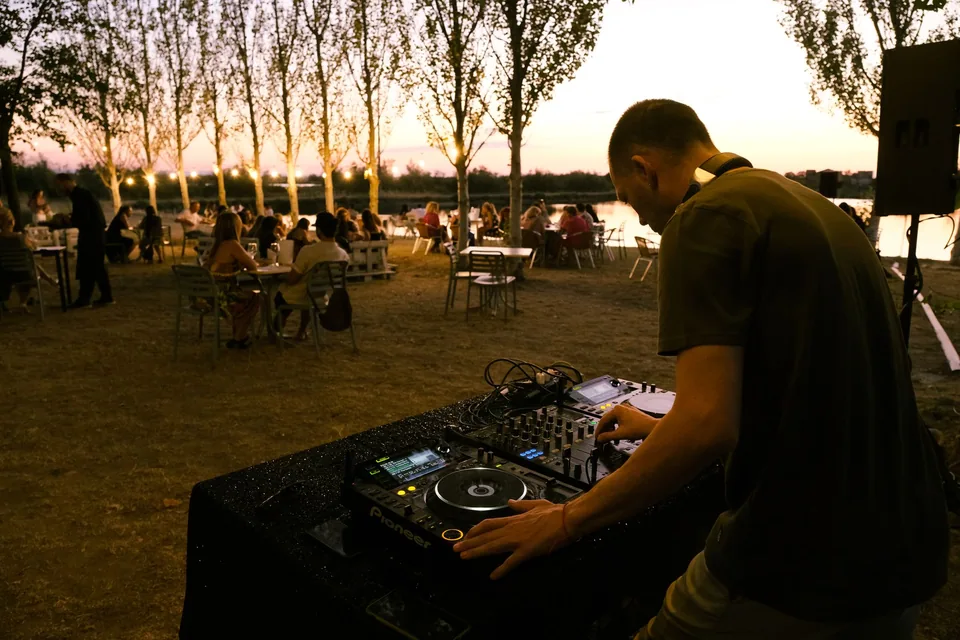 DJ tocando em mesa de som ao ar livre, à beira de um lago ao entardecer (foto de arquivo, não é do evento)