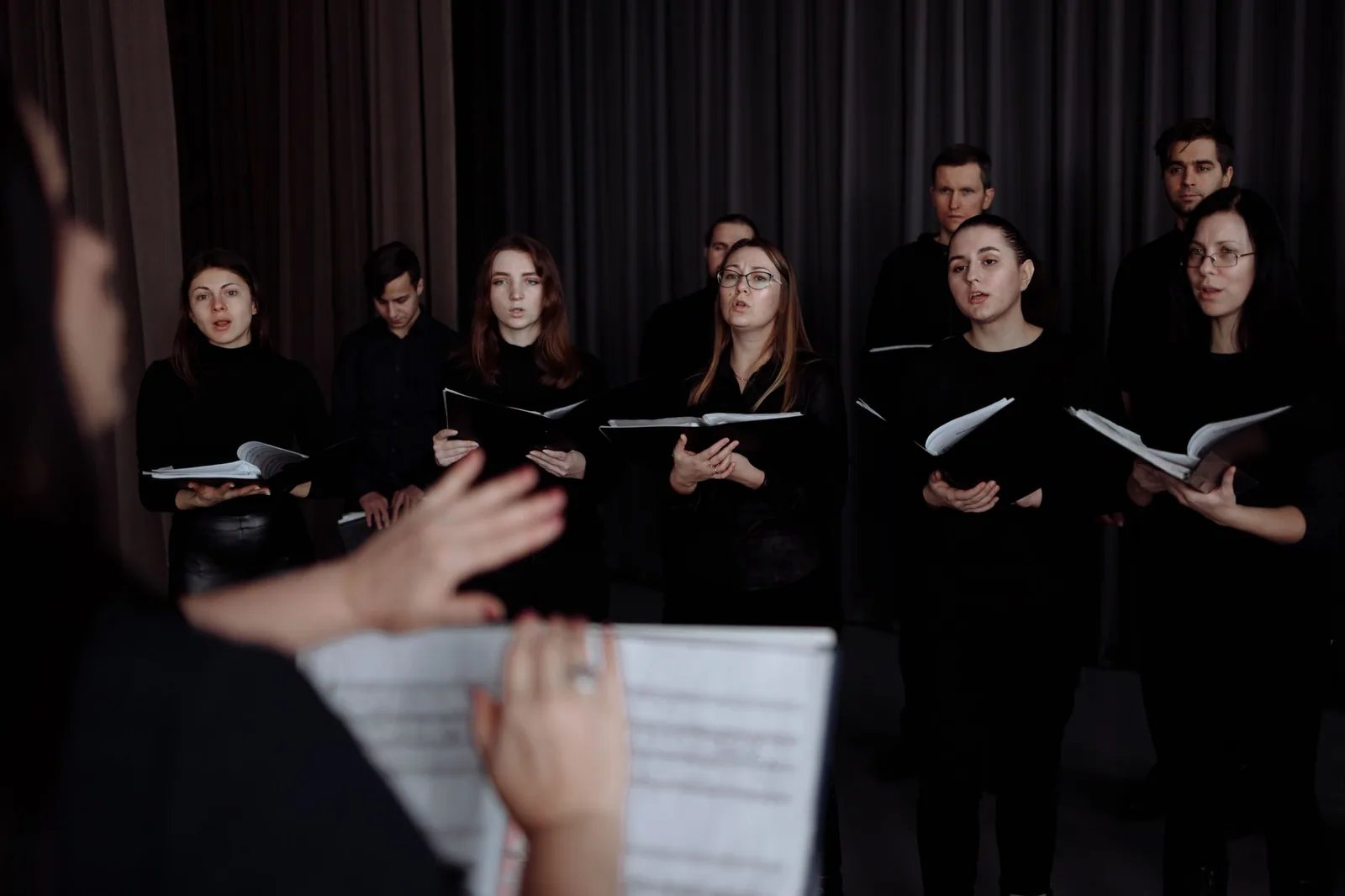 Coro de pé, de roupa preta e com pastas de partitura, olhando para a regente em primeiro plano, de costas, com as mãos levantadas