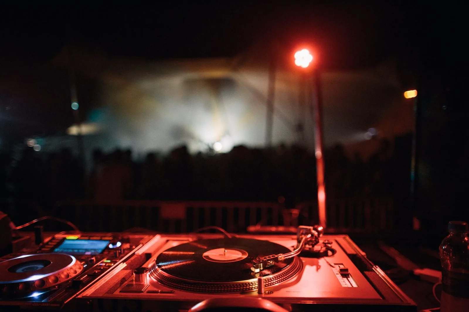 Toca-discos e controlador de DJ em primeiro plano, com palco e plateia desfocados ao fundo (foto ilustrativa)