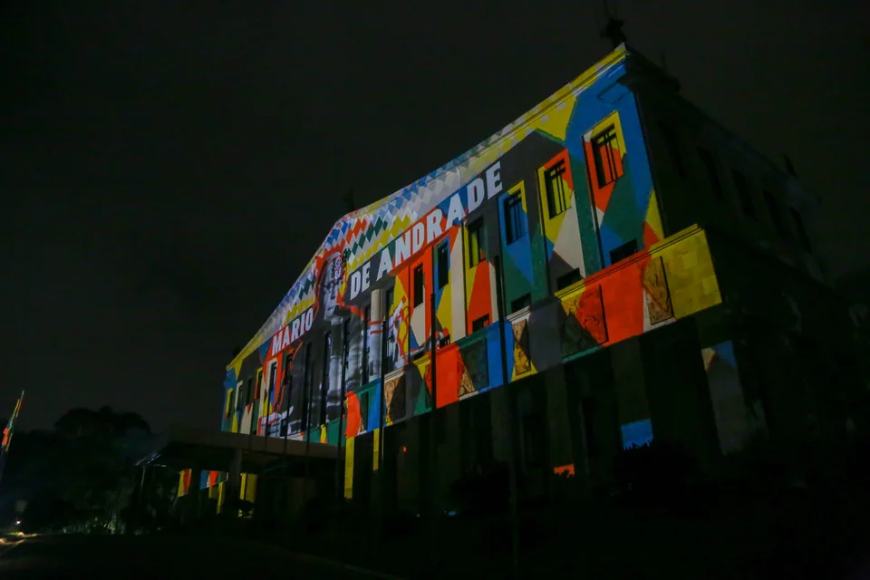 Fachada de um prédio em São Paulo iluminada por projeção de obras modernistas, com o nome Mário de Andrade, nos 100 anos da Semana de Arte Moderna