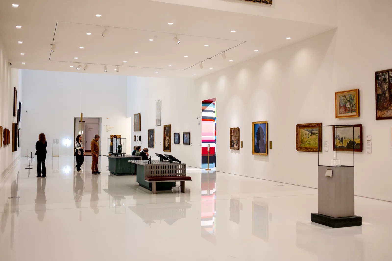 Sala de museu com paredes brancas, quadros emoldurados e visitantes circulando