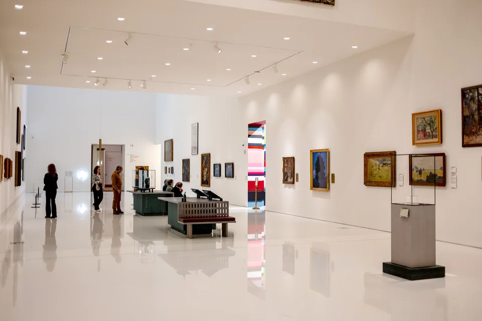 Sala de museu com paredes brancas, quadros emoldurados e visitantes circulando