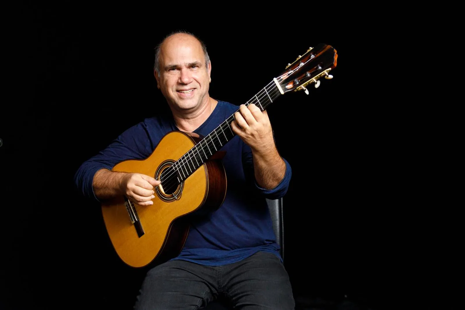 Nelson Faria sorrindo, sentado com um violão de cordas de nylon contra fundo preto (foto de divulgação sob licença CC BY 4.0)