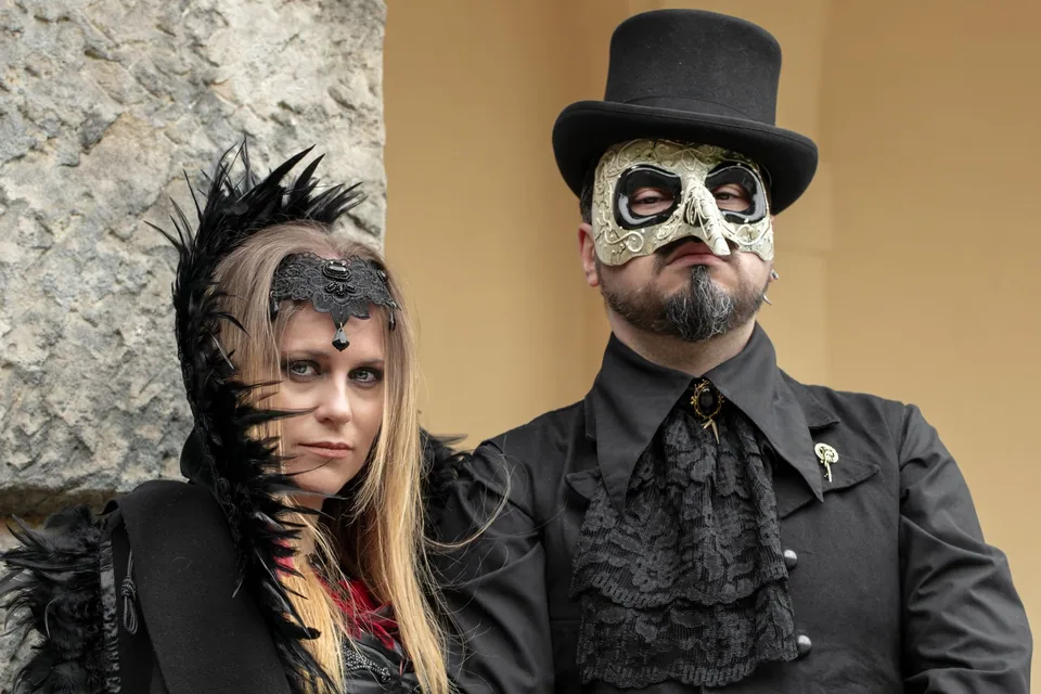 Casal em fantasia toda preta, ela com adereço de penas e renda na testa, ele com cartola e máscara clara de bico, em foto ilustrativa de festa de fantasia.