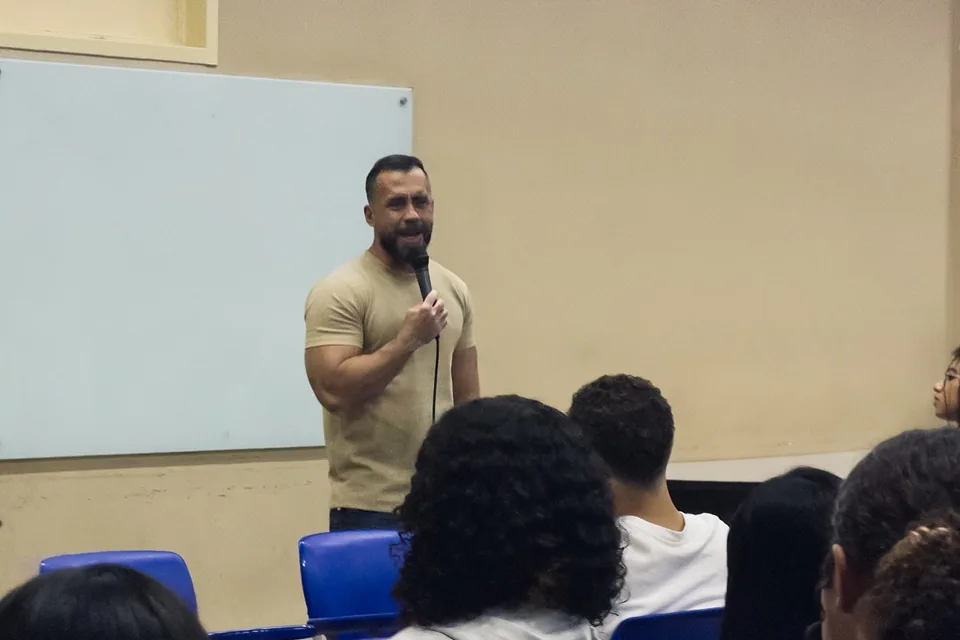 Professor de camiseta bege fala ao microfone diante de um quadro branco, em sala de aula com estudantes sentados de costas em cadeiras azuis.