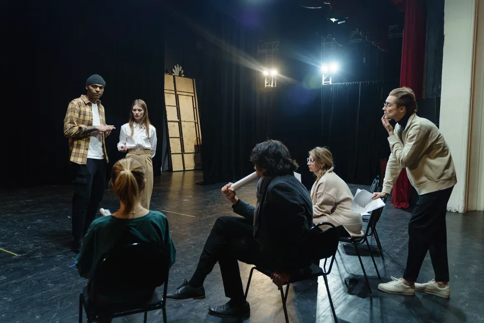 Grupo de atores em roda num palco de teatro, com dois em pé conversando e outros sentados em cadeiras