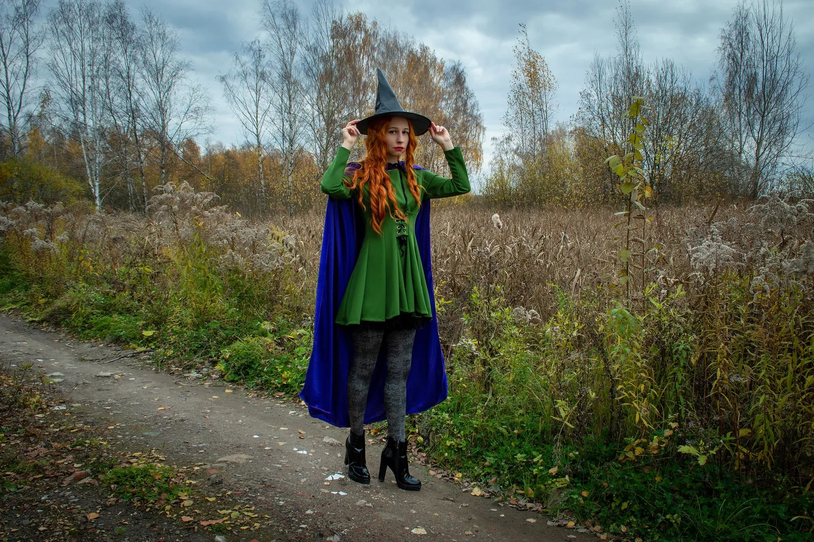 Mulher de vestido verde, capa azul e chapéu de bruxa em uma estrada de terra no outono
