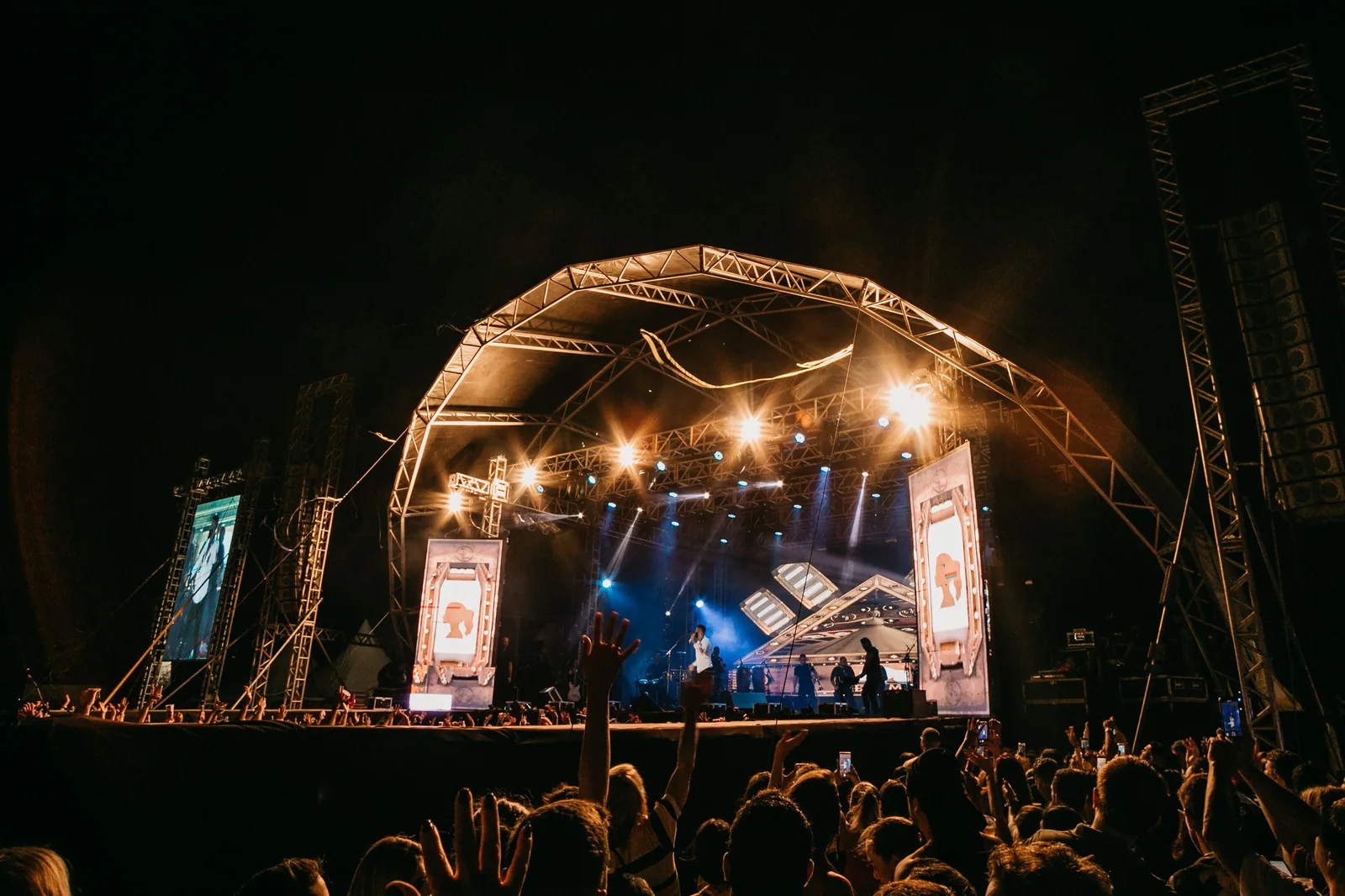 Palco de festival ao ar livre iluminado à noite, com público de braços erguidos na frente.