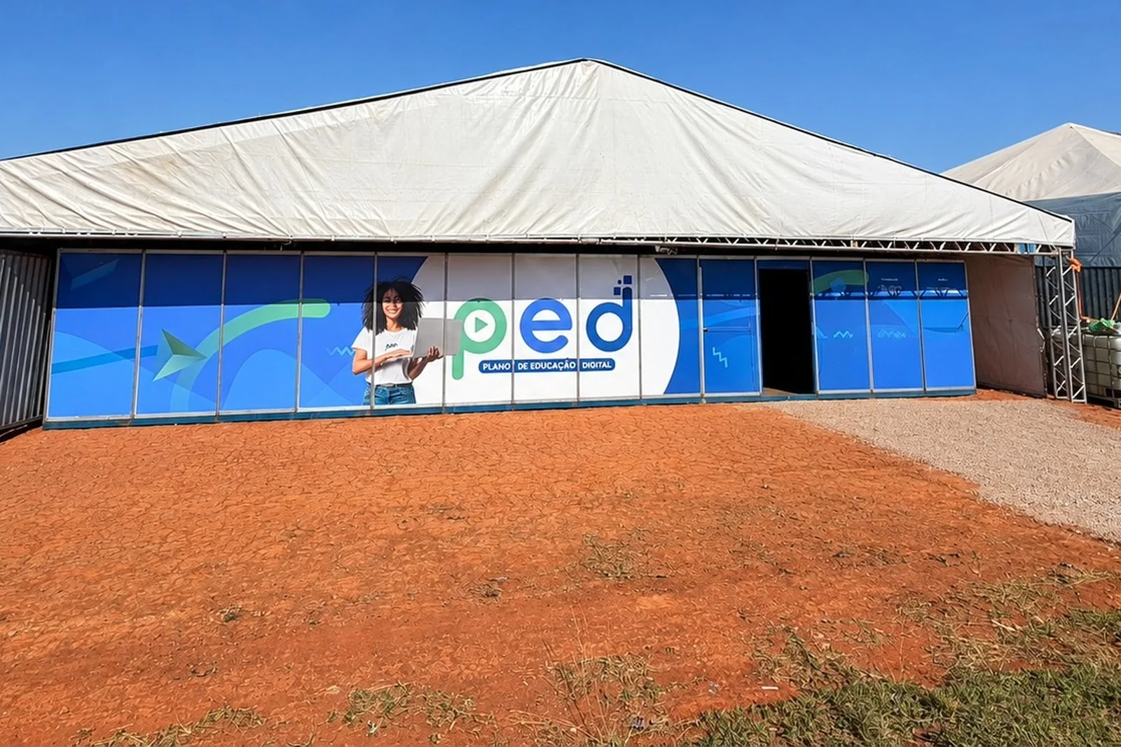 Fachada azul do PED, com a marca do projeto e a ilustração de uma jovem com notebook, sob galpão de lona branca
