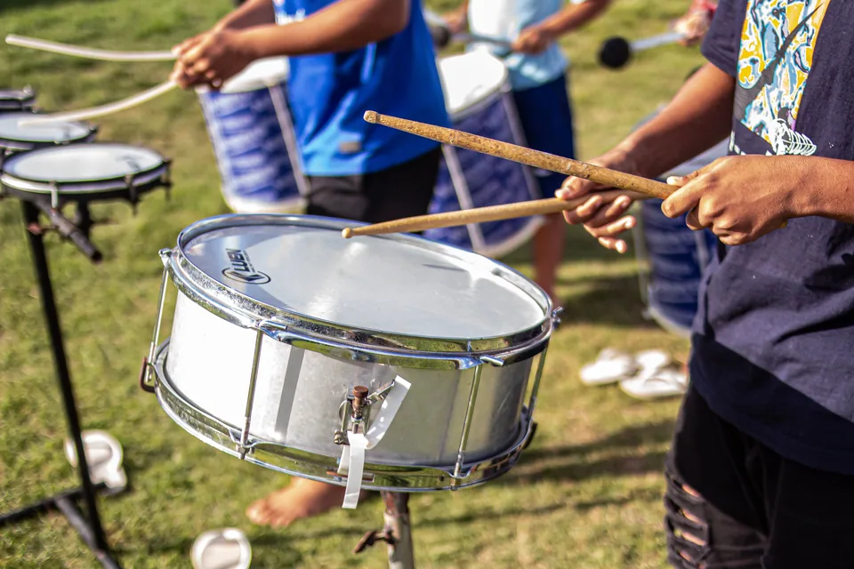 Mãos batendo com baquetas em uma caixa de percussão prateada, com outros percussionistas ao fundo em um gramado