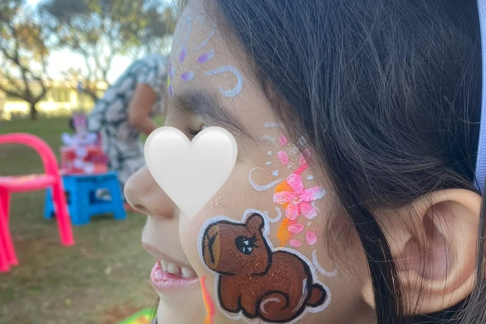 Menina de perfil com uma capivara e flores pintadas no rosto, num gramado com cadeirinhas cor-de-rosa ao fundo