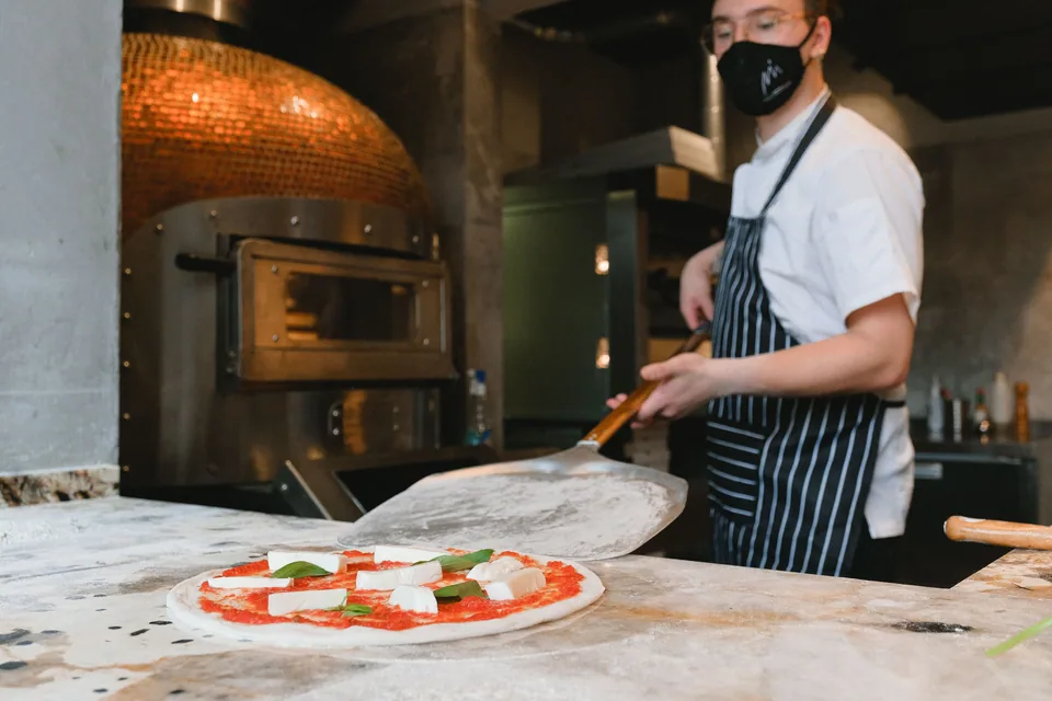 Pizzaiolo com pá de forno diante de forno a lenha, com uma pizza margherita crua na bancada
