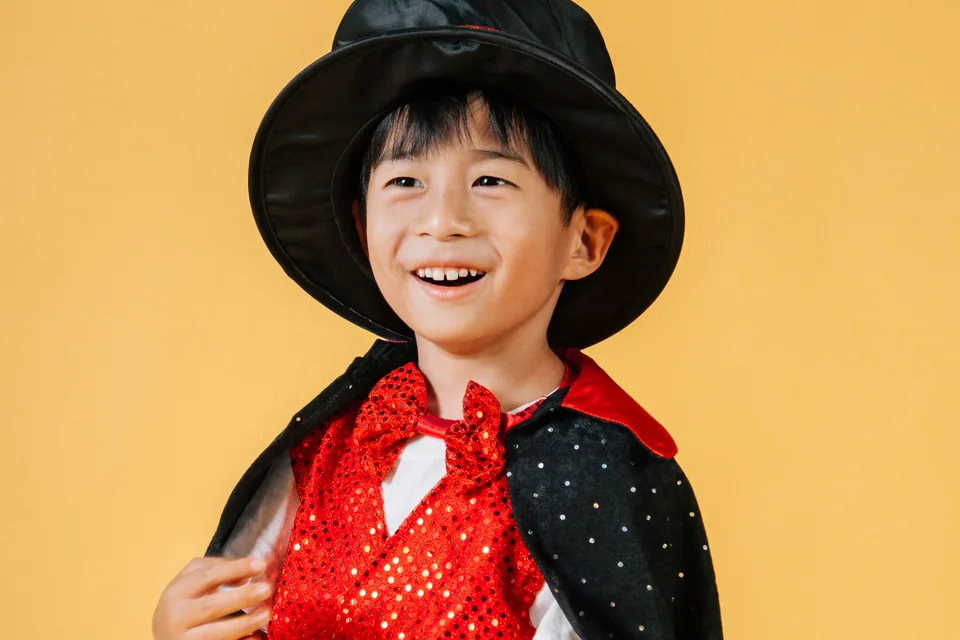 Criança fantasiada de mágico sorrindo, com cartola preta, colete vermelho e varinha na mão