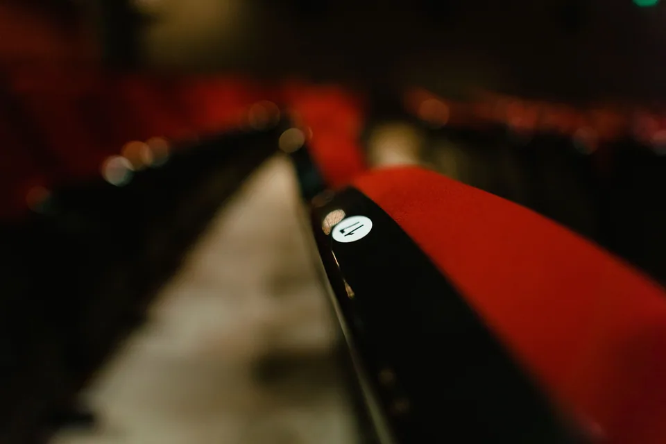 Fila de poltronas vermelhas de teatro em close, com a plateia vazia ao fundo