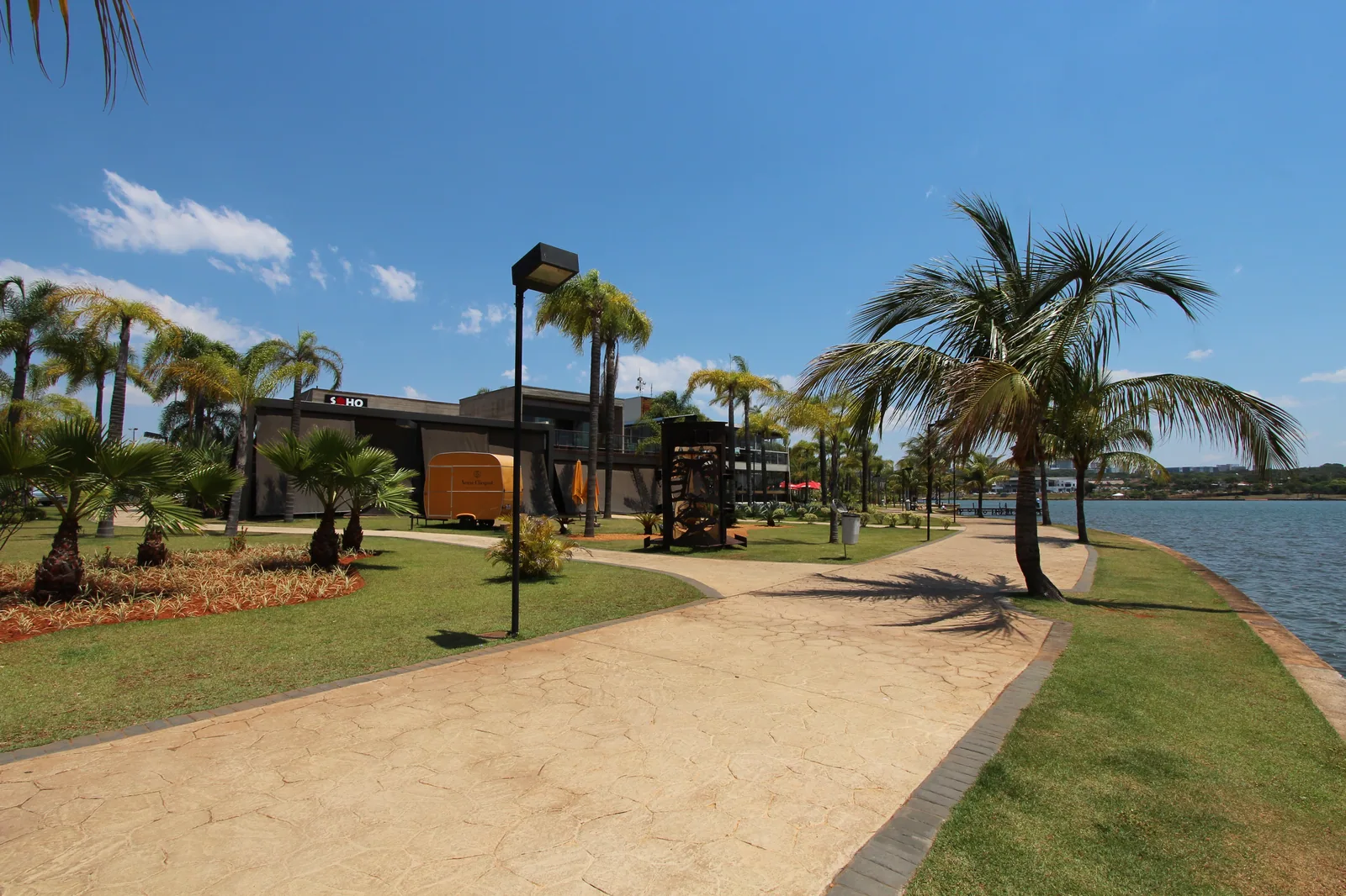 Calçadão do Pontão do Lago Sul entre palmeiras e o Lago Paranoá, com a fachada de um restaurante ao fundo