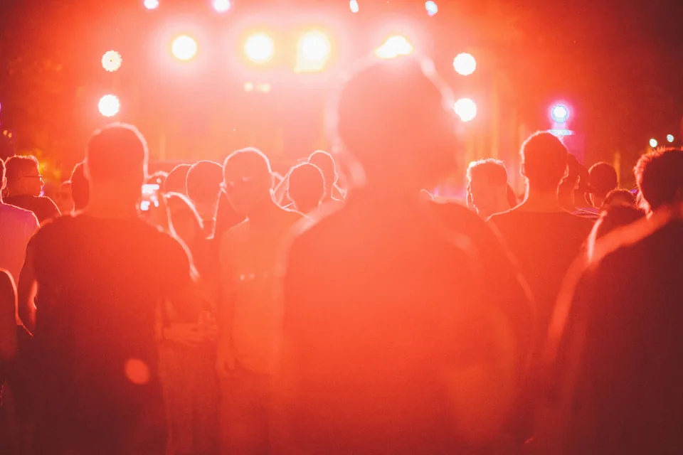 Publico de costas diante de um palco ao ar livre iluminado por luzes vermelhas, a noite