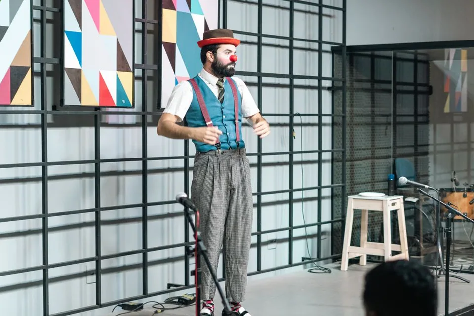 Palhaço de nariz vermelho, chapéu-coco e suspensório se apresenta em um palco, com instrumentos ao fundo