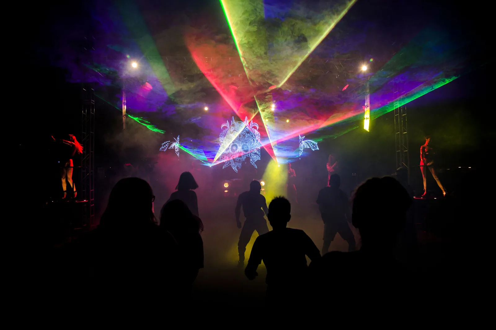 Pista de festa escura com feixes de laser coloridos, fumaça e silhuetas de pessoas dançando (foto ilustrativa de banco de imagens)