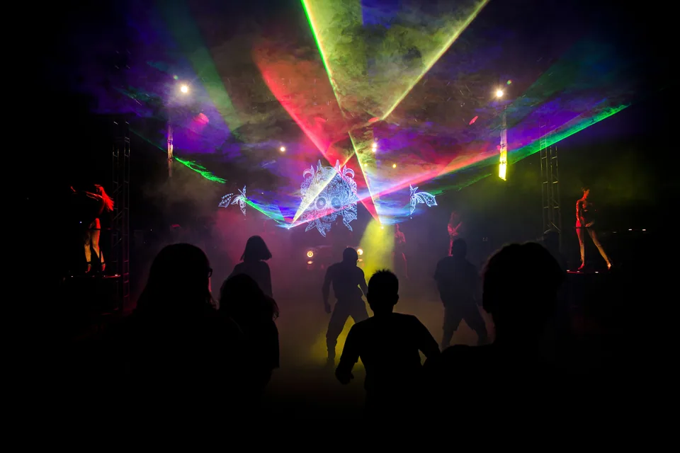 Pista de festa escura com feixes de laser coloridos, fumaça e silhuetas de pessoas dançando (foto ilustrativa de banco de imagens)