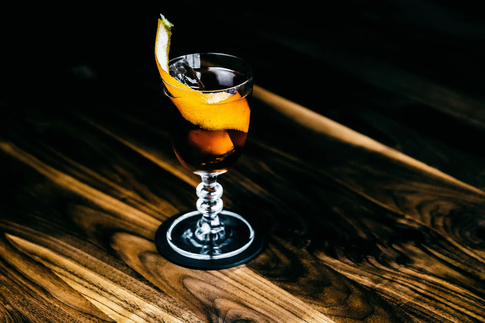 Coquetel âmbar em taça de pé alto com casca de laranja, sobre mesa de madeira escura