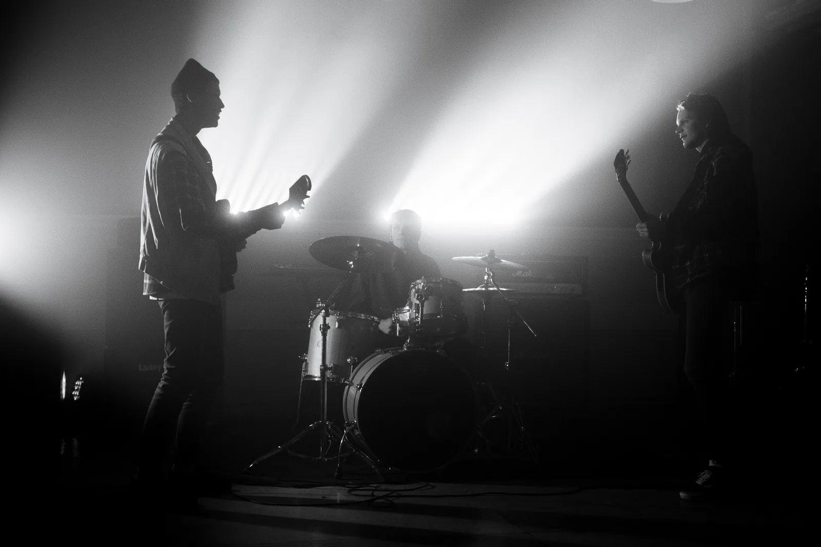 Três músicos em silhueta num palco com luz de contraluz: dois guitarristas e um baterista, em foto em preto e branco