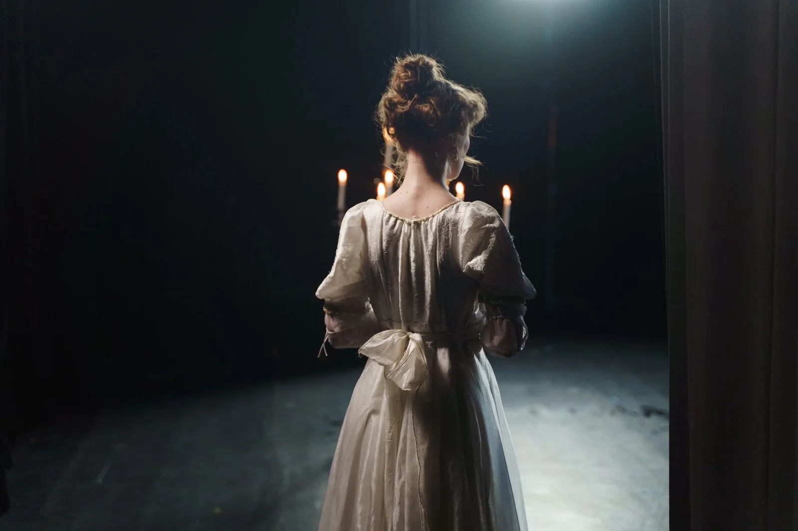 Atriz de vestido claro vista de costas no fundo de um palco escuro, com velas ao fundo, em foto ilustrativa
