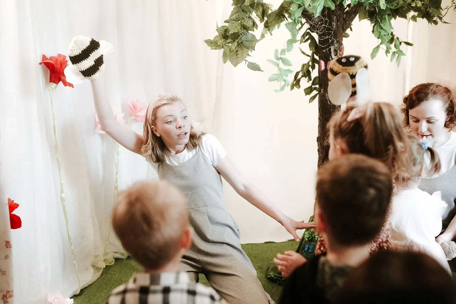 Atriz com fantoche de abelha encena peça infantil diante de crianças sentadas, em cenário de flores e árvore (foto ilustrativa)