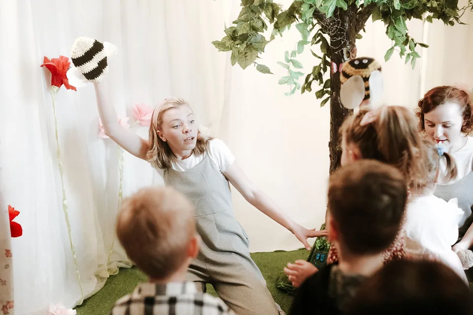 Atriz com fantoche de abelha encena peça infantil diante de crianças sentadas, em cenário de flores e árvore (foto ilustrativa)