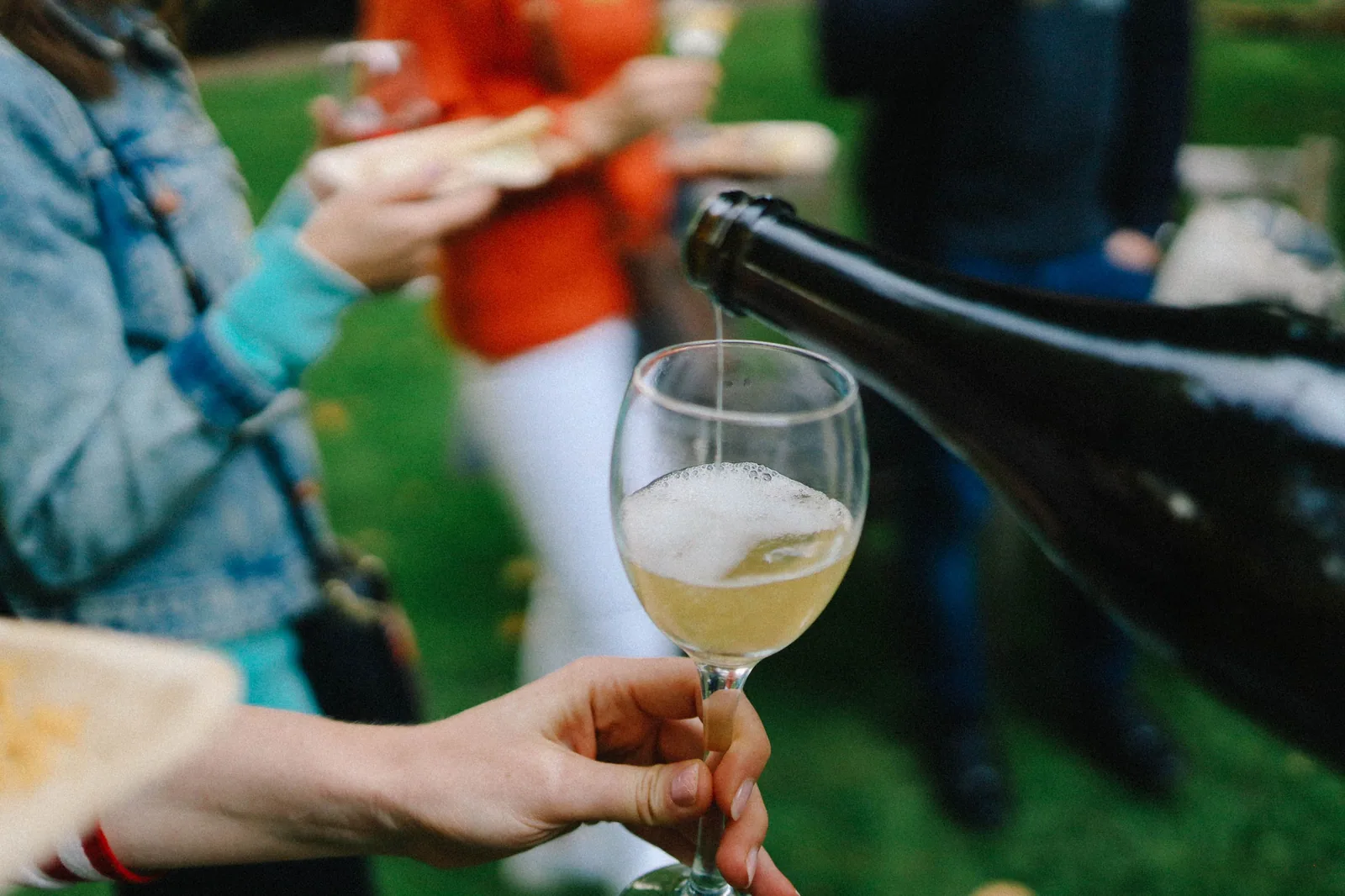 Taça de vinho branco sendo servida ao ar livre, com convidados ao fundo em evento de degustação