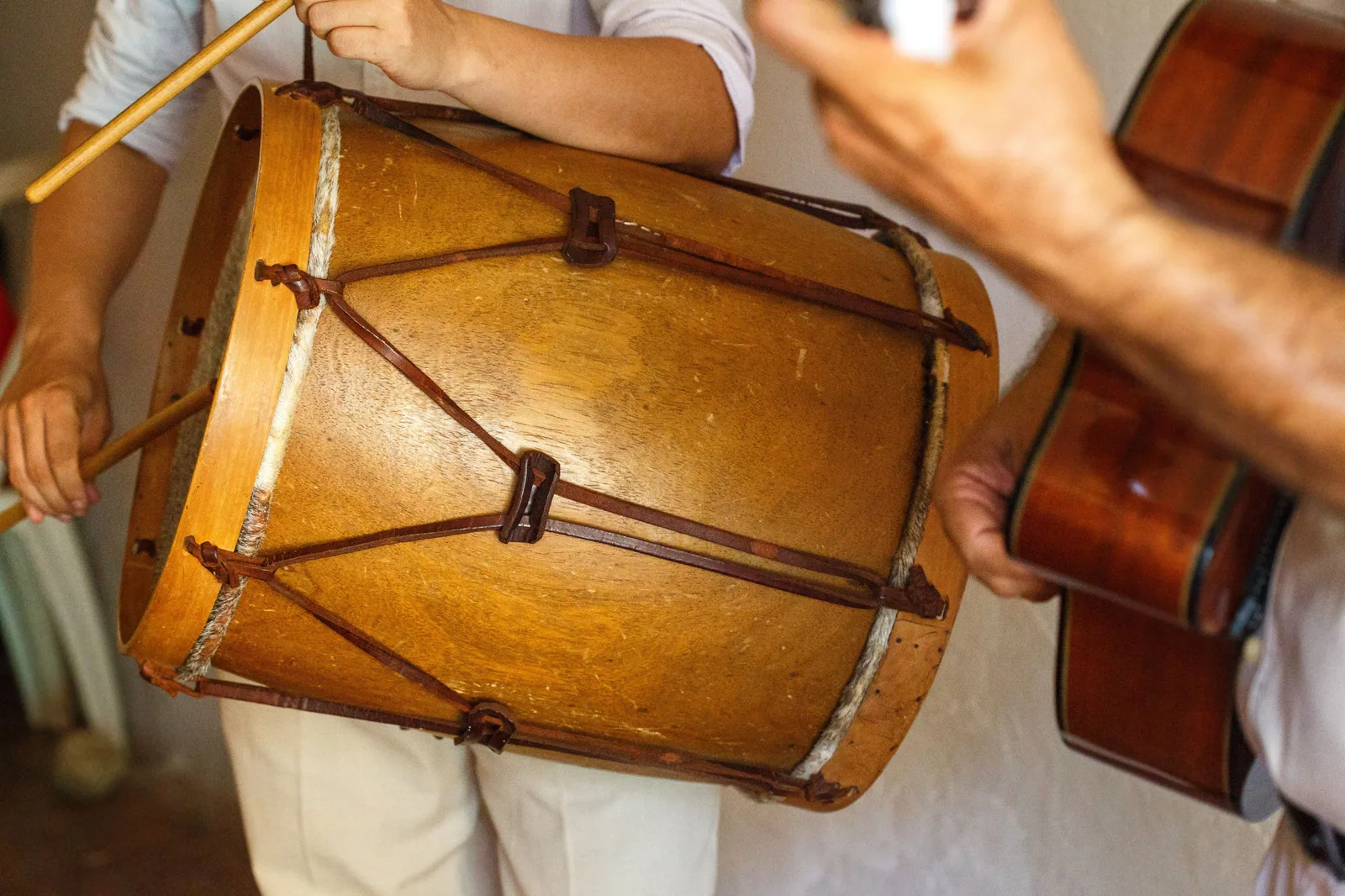 Mãos de músicos tocando um tambor de couro e um violão numa roda de samba