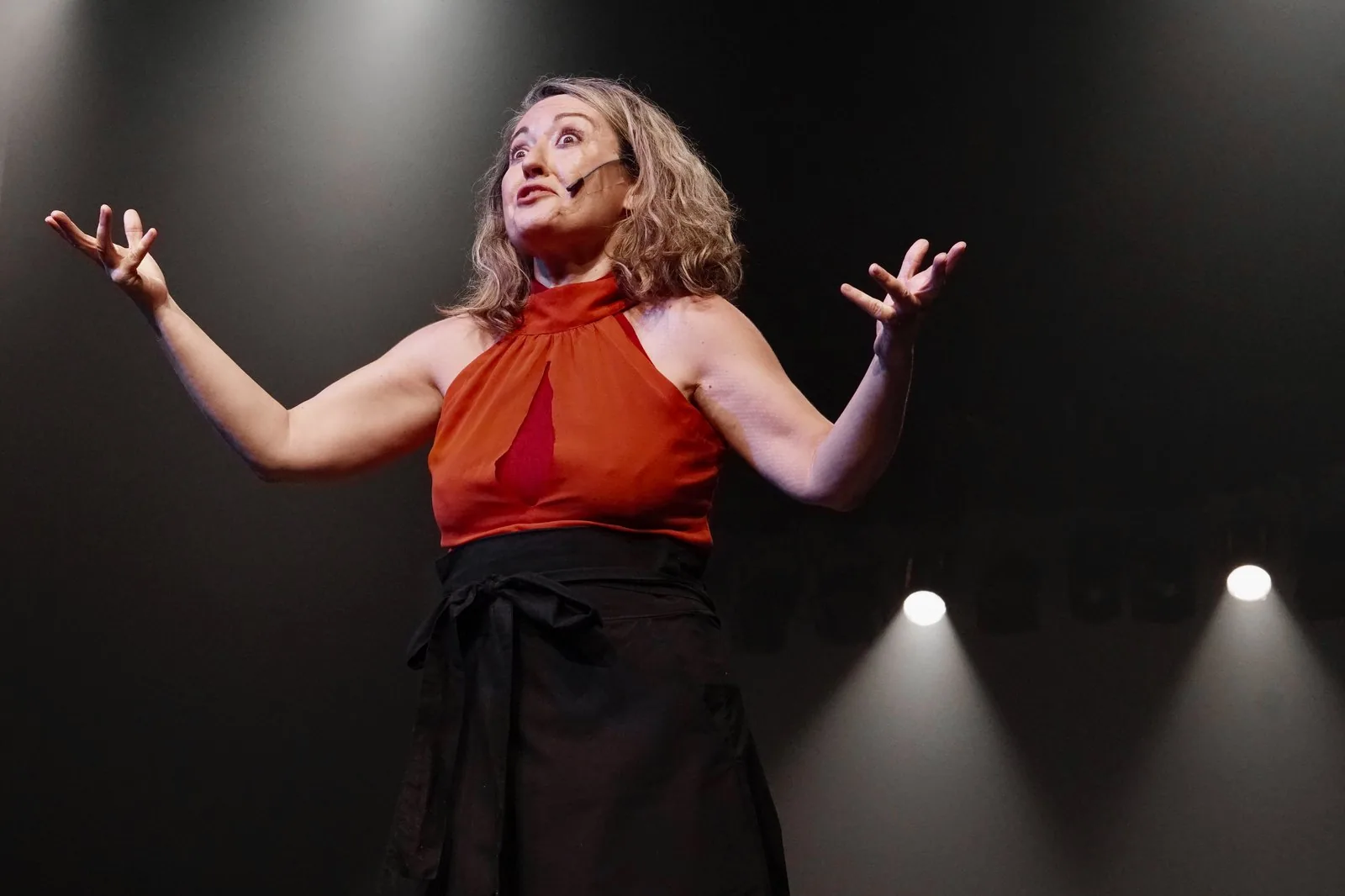 Atriz de blusa laranja abre os braços sob a luz do palco durante o espetáculo Senhora P