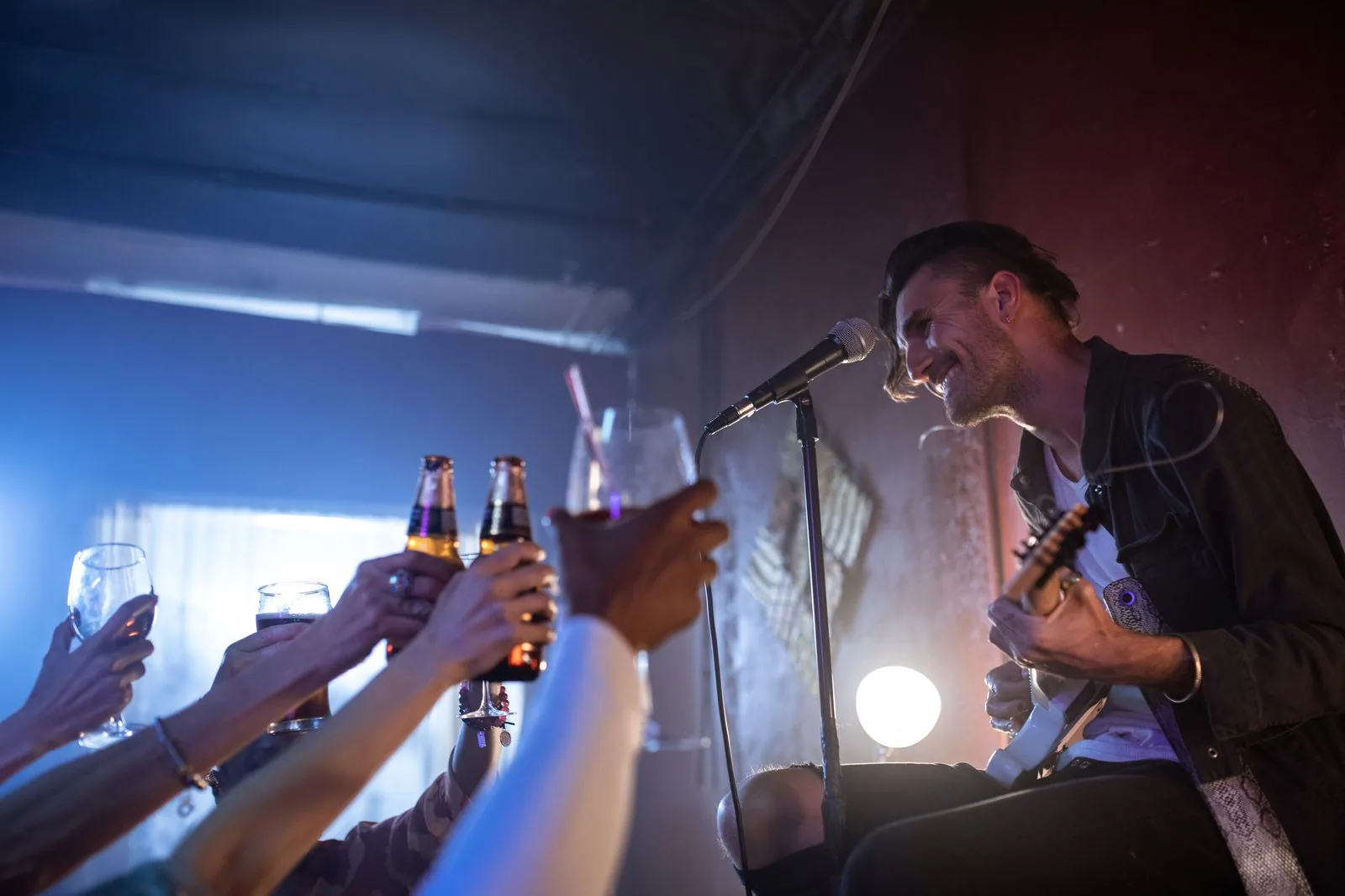 Cantor sorri ao microfone com a guitarra no colo enquanto o público ergue copos e garrafas para brindar