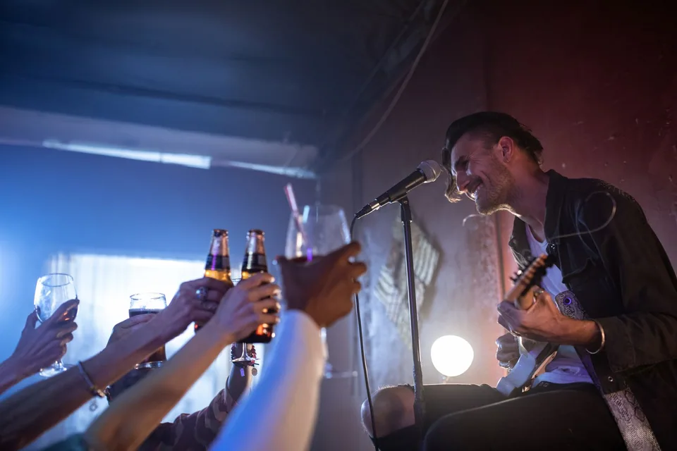 Cantor sorri ao microfone com a guitarra no colo enquanto o público ergue copos e garrafas para brindar