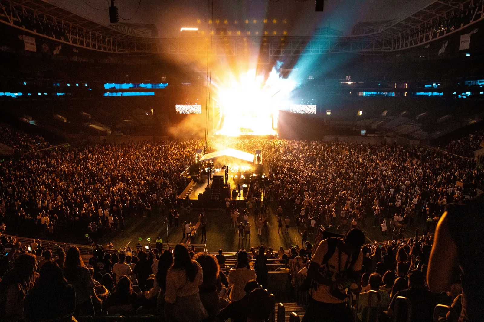 Estádio lotado durante show noturno, com palco iluminado ao centro e público em pé