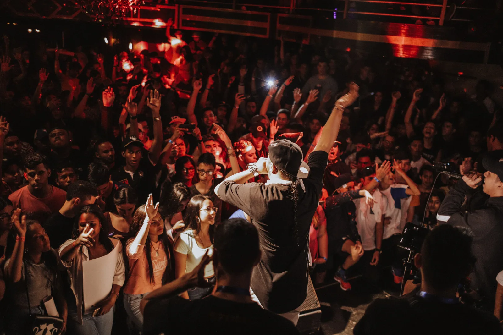 Público de mãos ao alto em show noturno, com o artista de costas no palco sob luz vermelha
