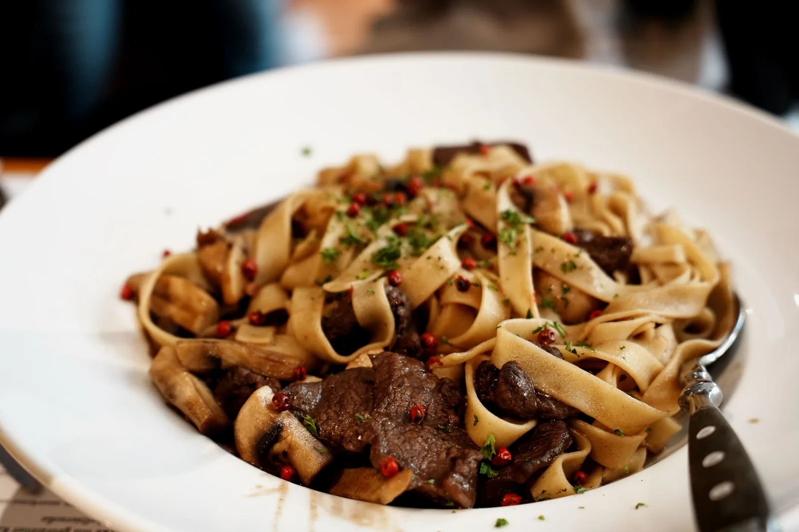 Prato fundo branco com tagliatelle, tiras de carne, cogumelos e pimenta rosa