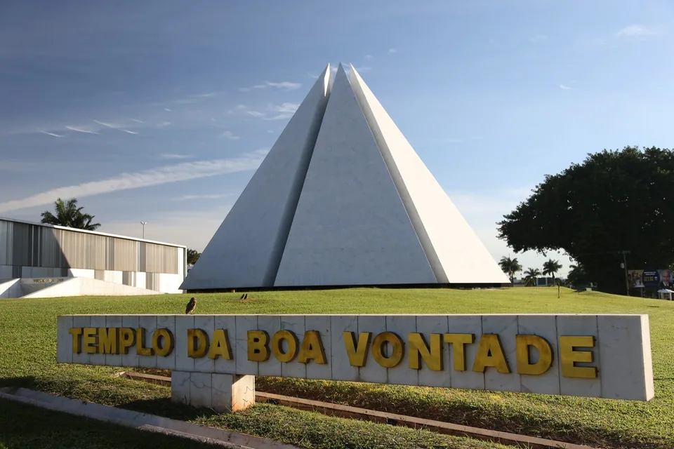 Pirâmide branca do Templo da Boa Vontade atrás do letreiro dourado com o nome do templo, num gramado, em Brasília.