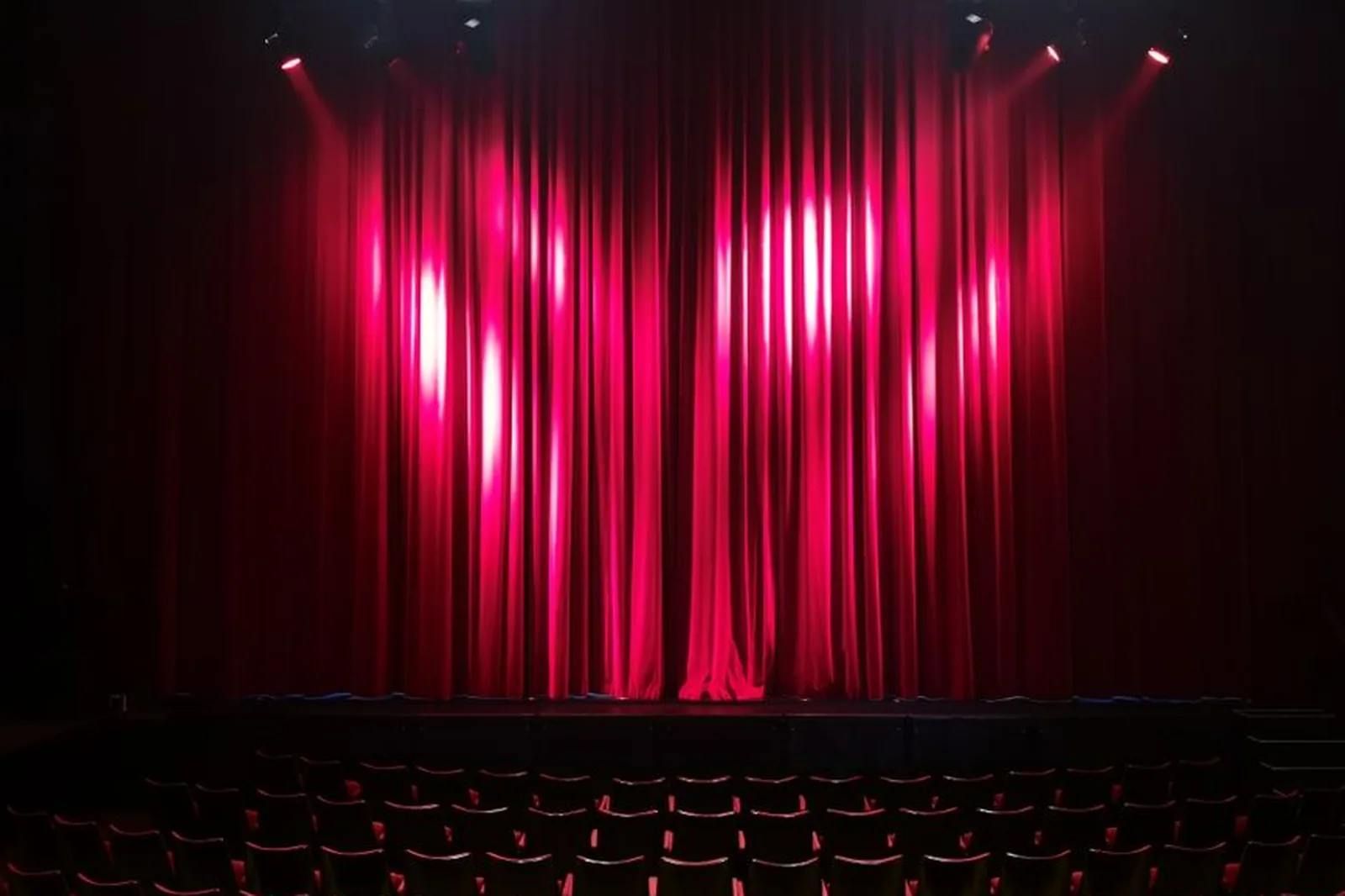 Cortina vermelha fechada iluminada por refletores sobre palco de teatro com poltronas vazias