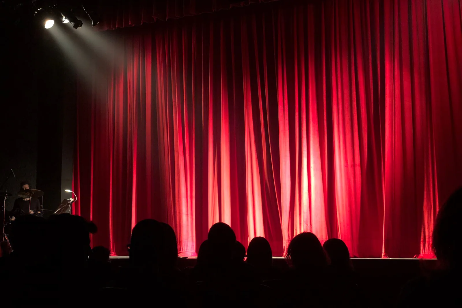 Plateia em silhueta diante de um palco com cortina vermelha iluminada por refletores