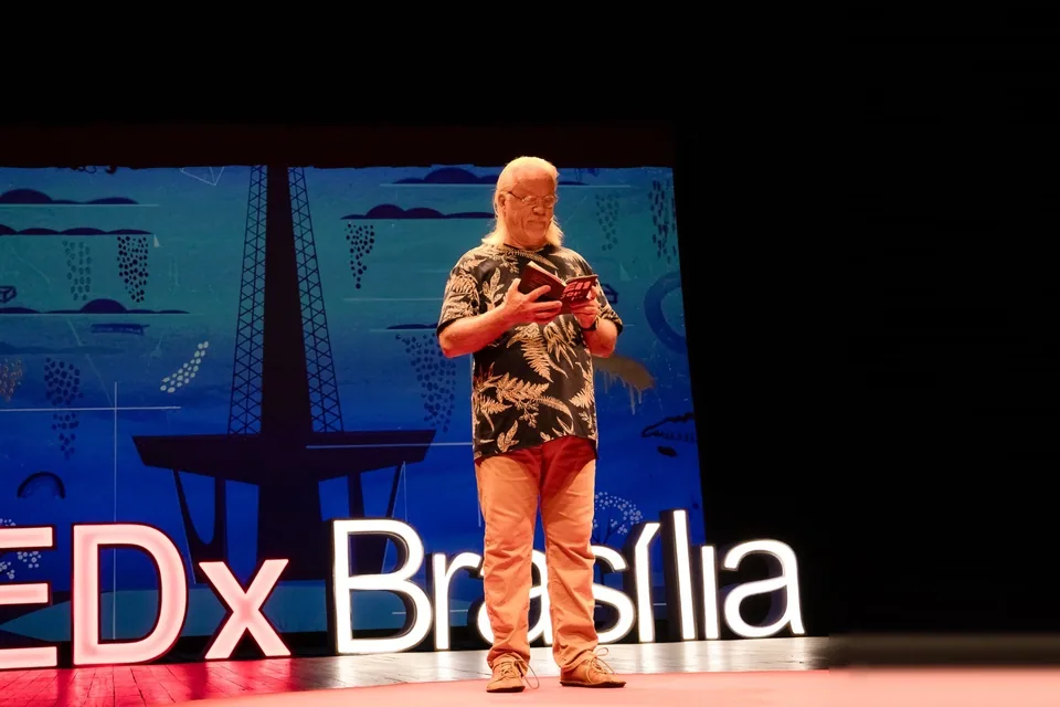 Palestrante lê um livro no palco do TEDxBrasília, com o letreiro luminoso e um painel azul com a Torre de TV ao fundo, em edição anterior