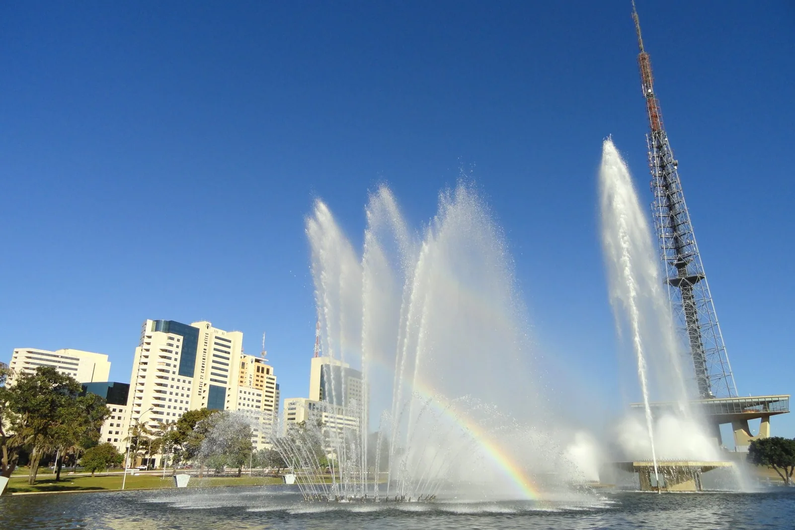 Fonte luminosa com jatos d'água e arco-íris em primeiro plano e a Torre de TV de Brasília à direita, sob céu azul