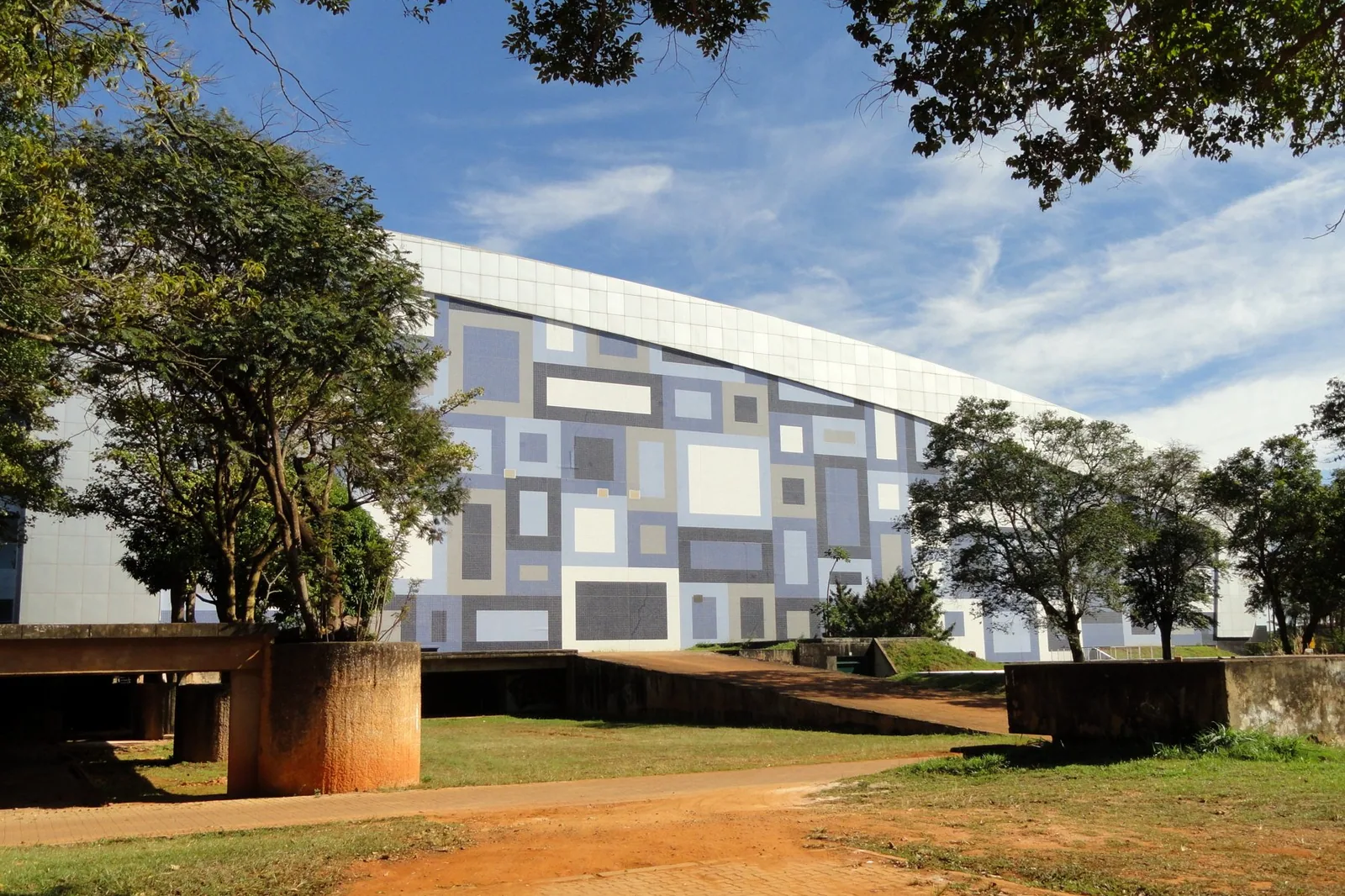 Fachada curva com painel de azulejos em tons de azul e cinza do Centro de Convenções Ulysses Guimarães, em Brasília, vista entre árvores