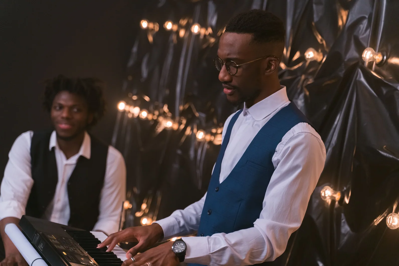 Pianista de colete azul toca teclado ao lado de outro músico em palco com luzes