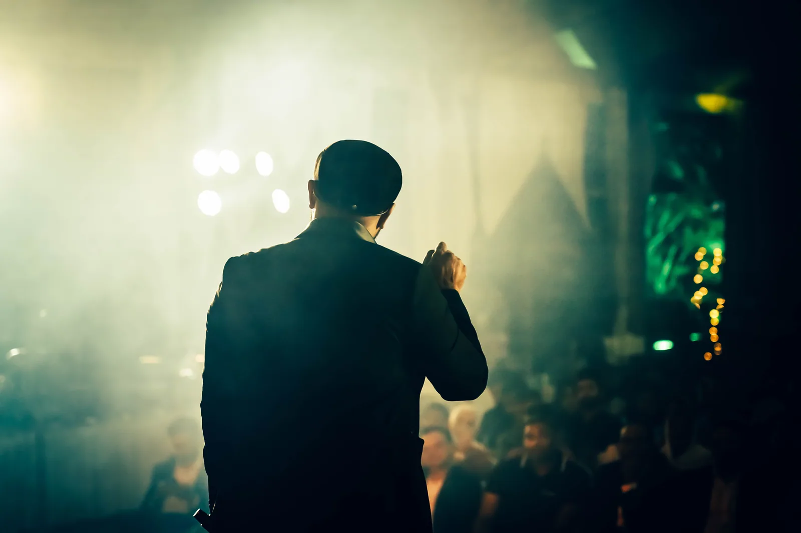 Humorista de costas, com o microfone na mão, diante da plateia em um palco com fumaça e luzes