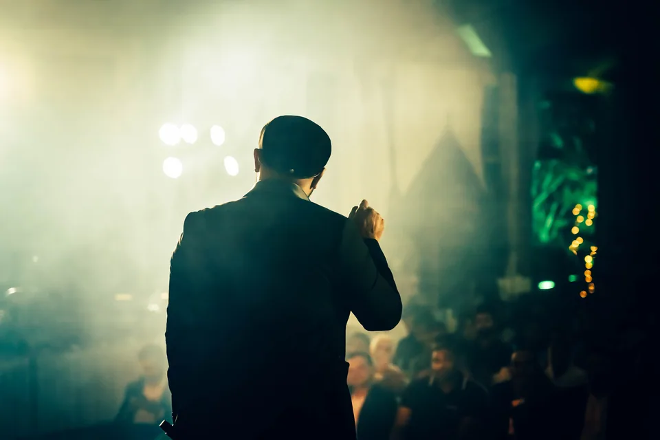 Humorista de costas, com o microfone na mão, diante da plateia em um palco com fumaça e luzes