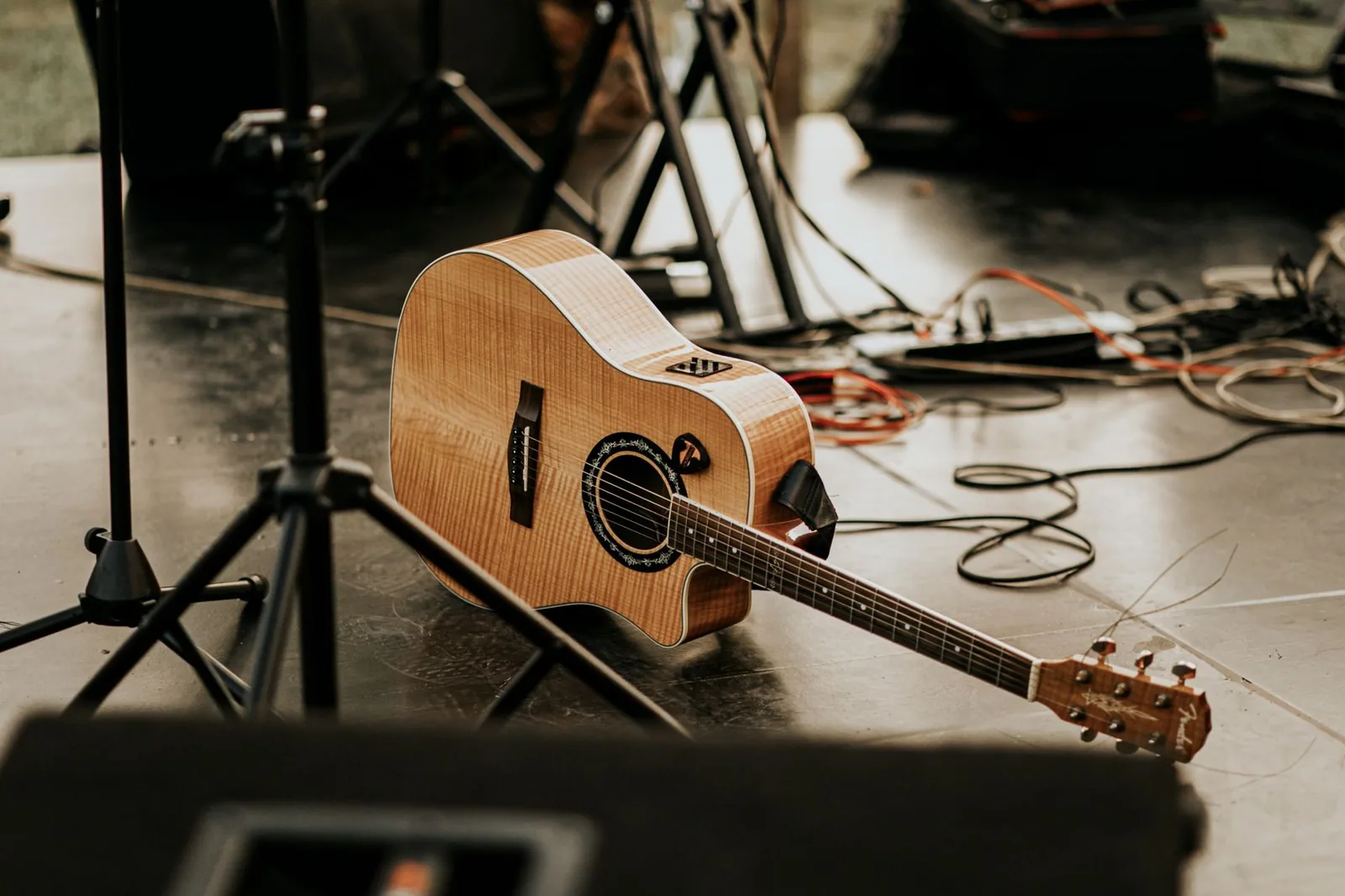 Violão acústico apoiado no chão de um palco, entre pedestais e cabos de som